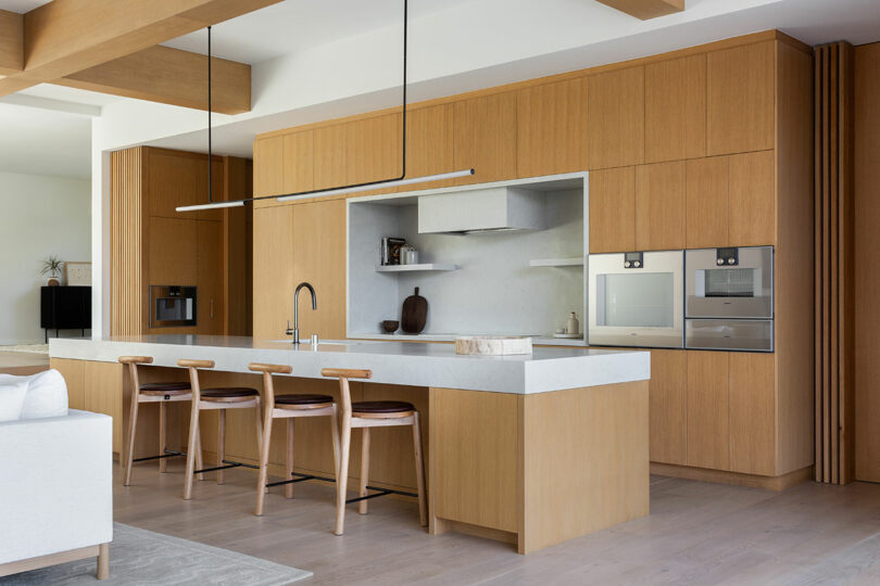 La cocina moderna cuenta con gabinetes de madera clara, encimeras blancas, electrodomésticos integrados, una gran isla con cuatro bancos de madera, estantes abiertos y una decoración minimalista.