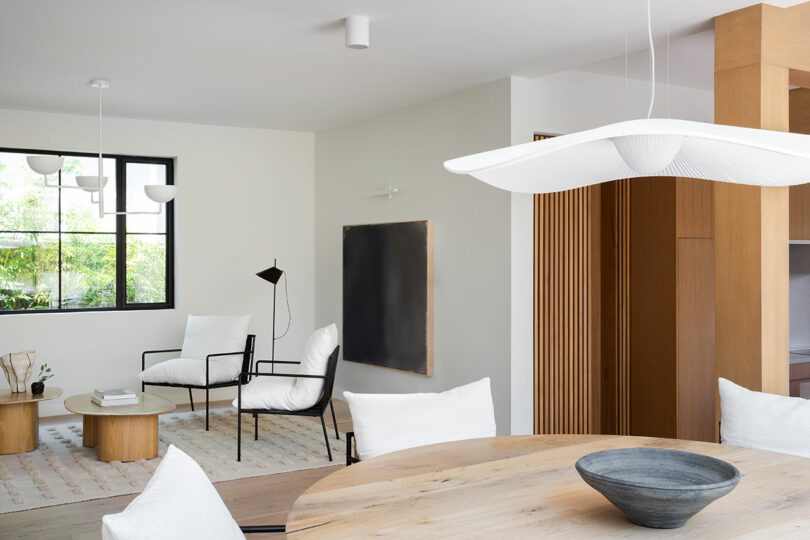 La moderna sala de estar y comedor cuenta con muebles de madera clara, cojines blancos, una mesa de comedor redonda, grandes ventanales, una decoración minimalista y tonos neutros.