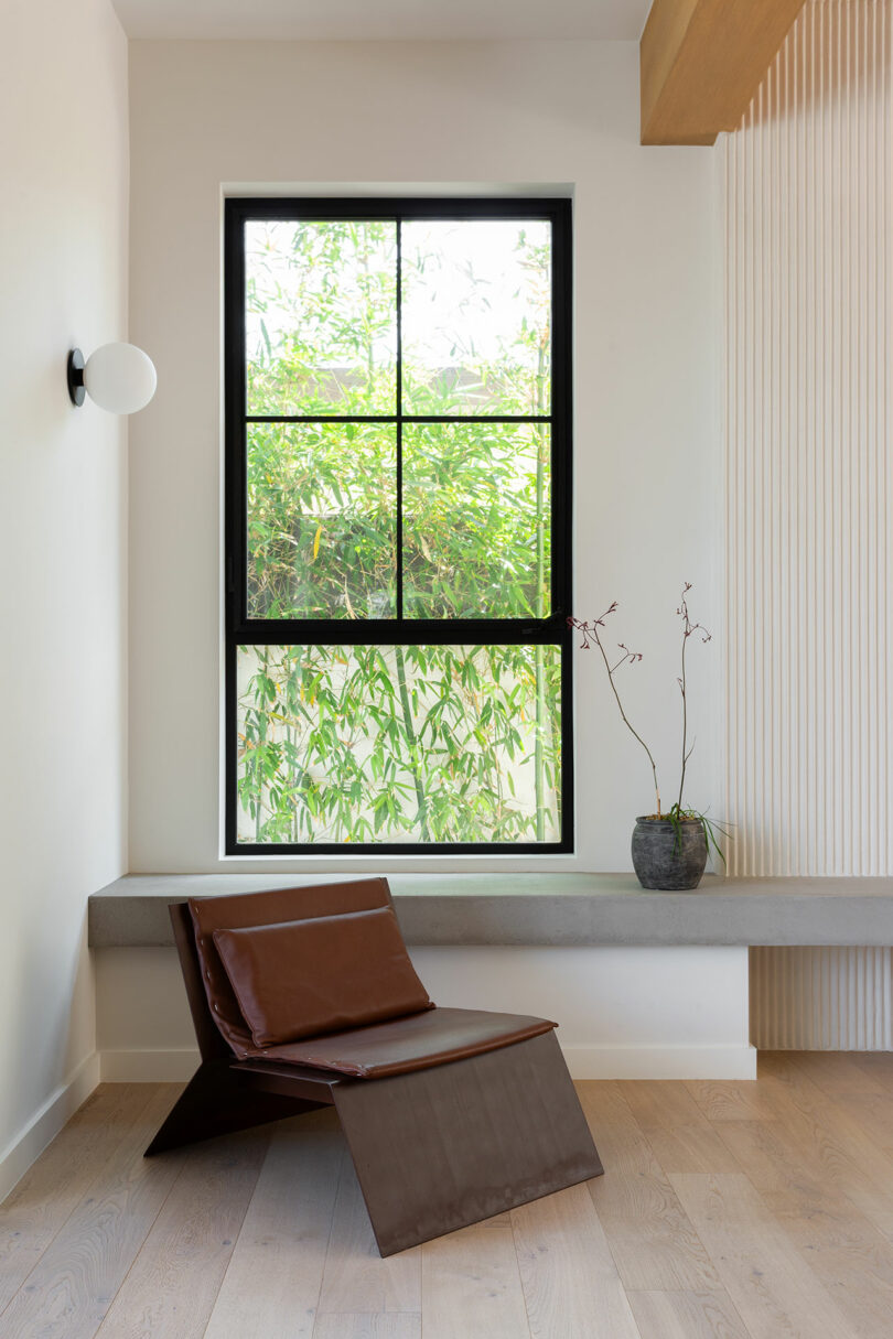 Una habitación minimalista con modernas sillas marrones, orquídeas en macetas sobre una repisa de hormigón, grandes ventanales y bambú visible en el exterior.