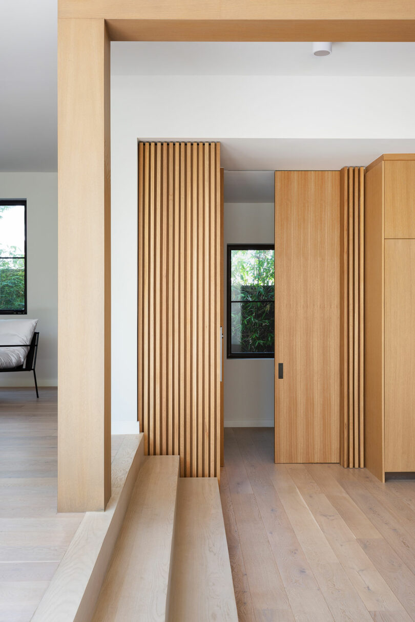 Un interior minimalista con chapas de madera clara, paneles de listones de madera verticales, suelos de dos niveles y una ventana que da a la vegetación exterior. Parte de la cama se puede ver en la habitación contigua.