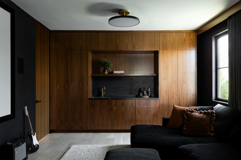 Salón moderno con armarios de madera oscura, lavabo empotrado, estantes decorativos, sofá seccional negro y ventana con cortinas grises.