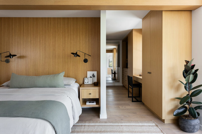 Dormitorio moderno con paredes de paneles de madera, cama ordenada, lámpara de pared, escritorio empotrado y maceta sobre suelo de madera clara.