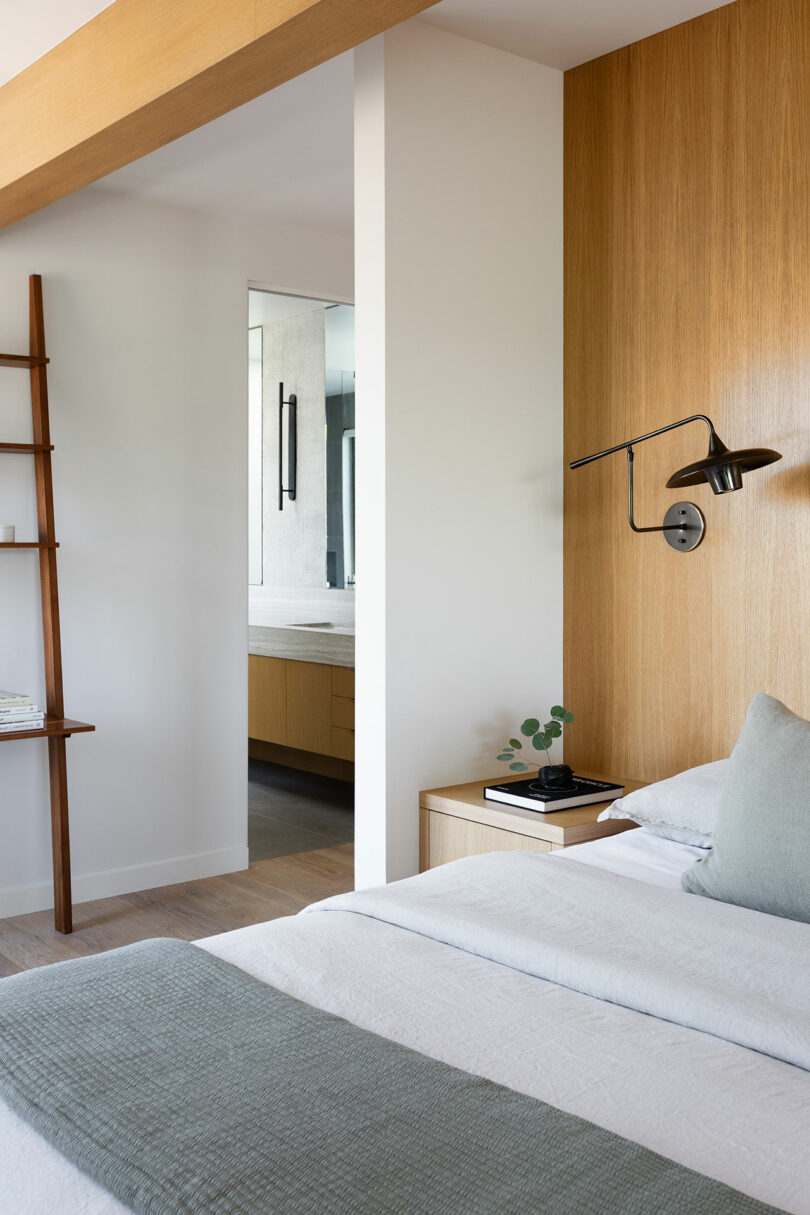 Dormitorio moderno con decoración de madera, cama cuidada, lámpara de pared y vistas a un baño minimalista con armarios luminosos.