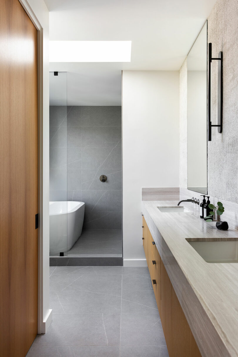 El moderno baño cuenta con gabinetes de madera clara, un espejo grande, un lavabo rectangular, una ducha con puerta de vidrio y una bañera independiente. Se utilizan tonos neutros de gris y blanco en todas partes.