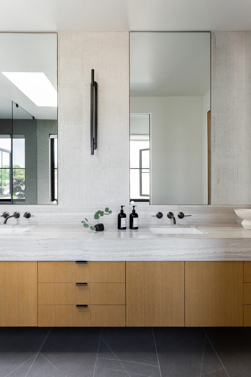 El moderno baño cuenta con un lavabo doble, un tocador de madera clara, accesorios negros, dos espejos grandes y una decoración minimalista que incluye un dispensador de jabón y ramitas en un jarrón.