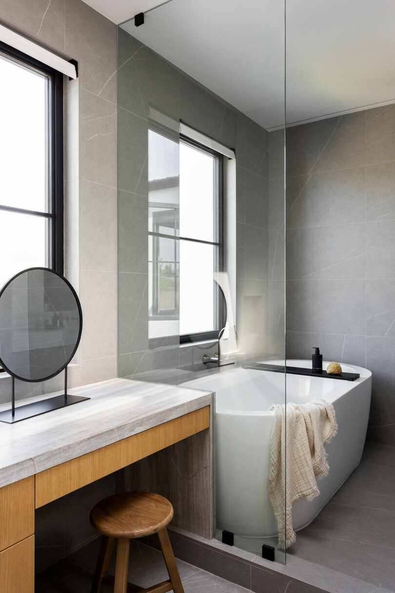 El moderno baño cuenta con una bañera independiente, toallas con cortinas, un tocador de madera con espejo redondo, un taburete de madera y una gran ventana que deja entrar luz natural.