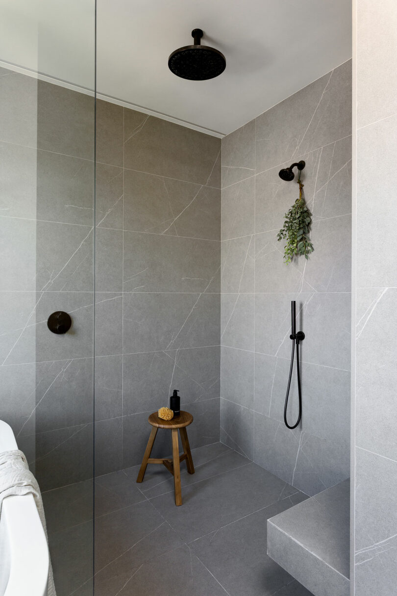 Cuarto de baño moderno con azulejos grises de gran formato, ducha a ras de suelo, accesorios negros, un pequeño banco de madera con esponjas y botellas, plantas verdes colgadas en la pared.