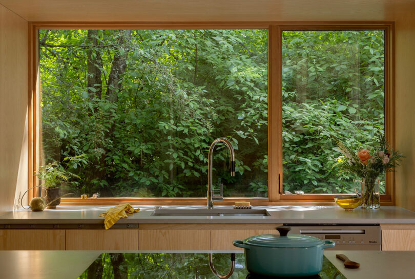 A modern kitchen sink with a large window overlooks dense green foliage; a turquoise pot and flowers sit on the countertop.