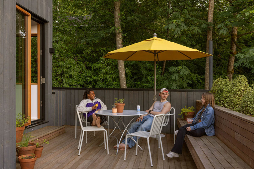Three people sit and talk around a round table on a wooden patio with potted plants and a yellow umbrella, surrounded by trees.