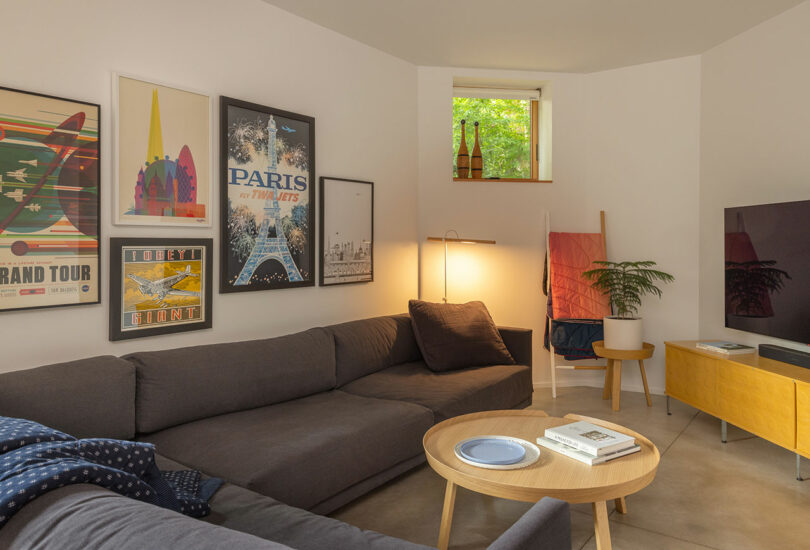 Modern living room with gray sectional sofa, colorful travel posters on the wall, a small window, indoor plant, TV, wooden furniture, and books on a round coffee table.