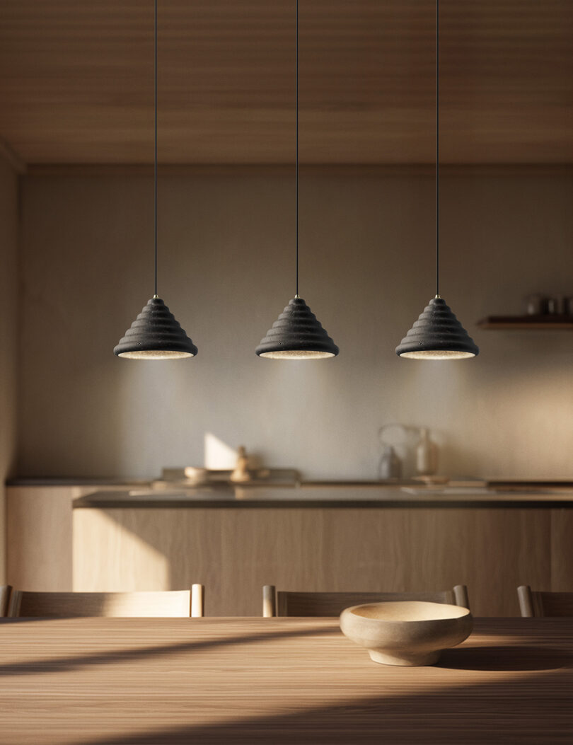 Tres luces colgantes negras cuelgan sobre una mesa de comedor de madera con un recipiente redondo en una cocina minimalista con acabados en madera clara.