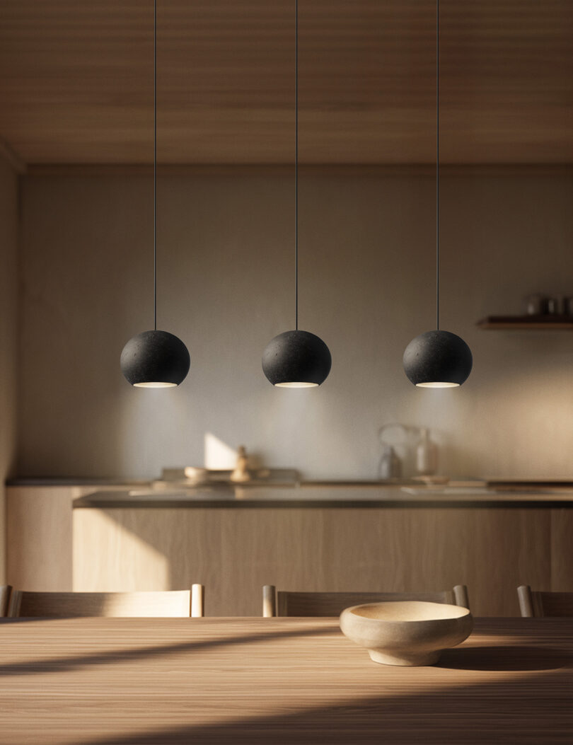 Tres luces colgantes negras cuelgan sobre una mesa de comedor de madera con tazones de madera, y la cocina está equipada con gabinetes y estantes de madera clara.