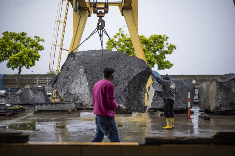 Dos trabajadores guían una gran roca suspendida de una grúa en un sitio industrial al aire libre con árboles y suelo húmedo.