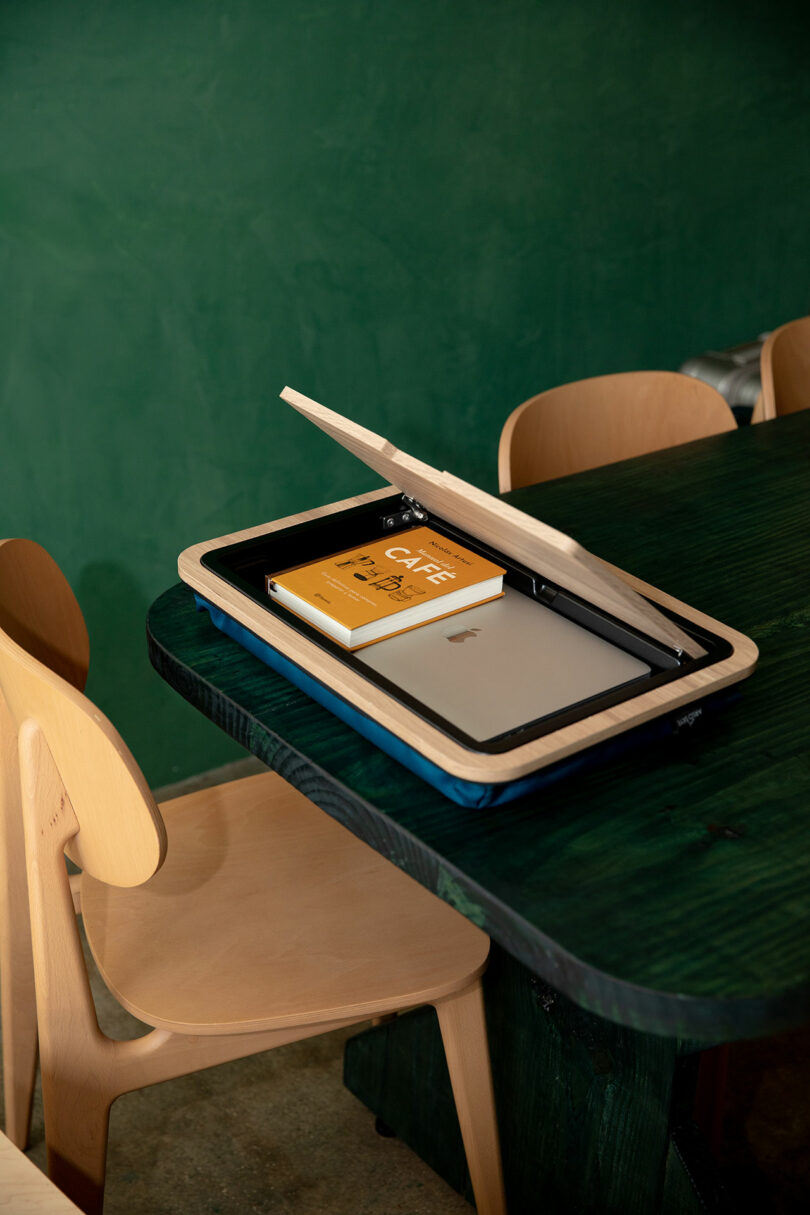 Una caja de madera con la tapa parcialmente abierta, que contiene un portátil plateado cerrado y un libro amarillo, se encuentra sobre una mesa verde oscuro junto a una silla de madera clara.