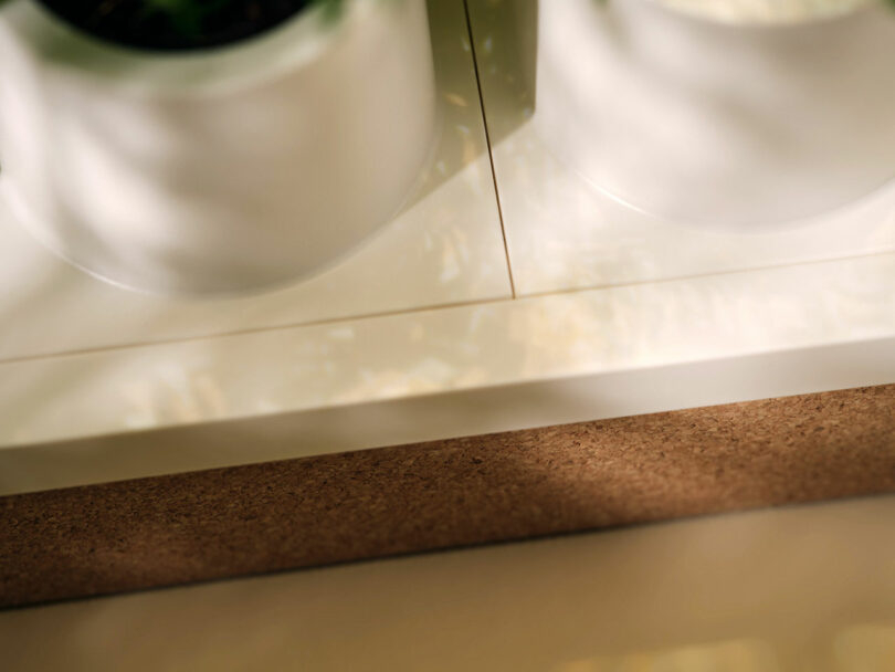 Dos lavabos de cerámica blanca se colocan sobre una superficie blanca junto a un tablero de corcho marrón con un patrón de luces y sombras visibles en la superficie.