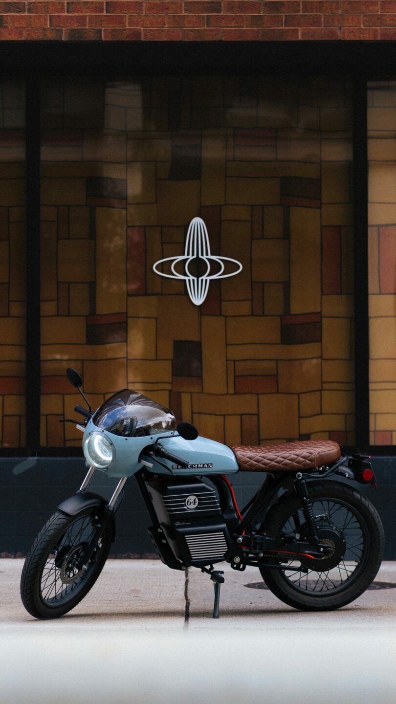 Una motocicleta eléctrica moderna con un asiento de cuero marrón está estacionada frente a una vidriera con un diseño geométrico y un logotipo circular.