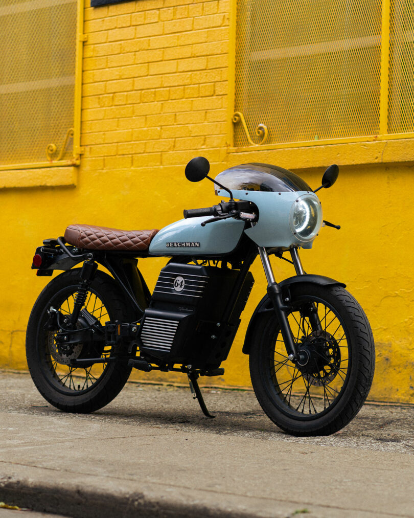 Una motocicleta eléctrica de estilo retro con asiento de cuero marrón está estacionada en la acera húmeda frente a una pared de ladrillos de color amarillo brillante.