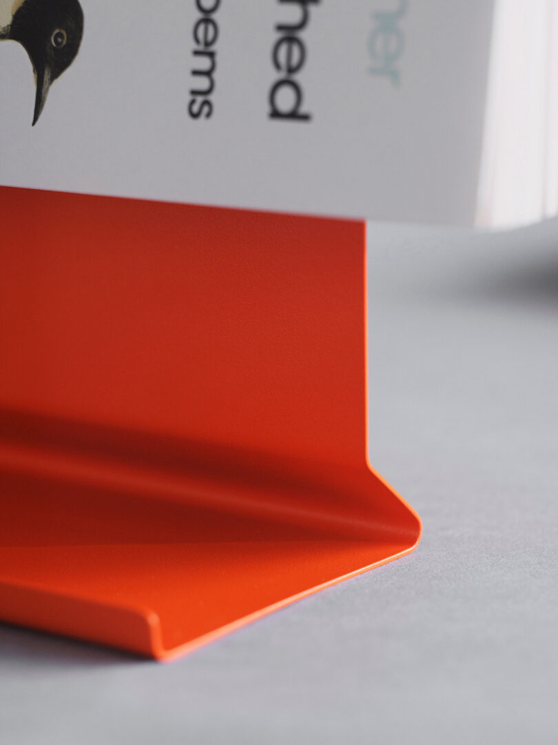 Un primer plano de un libro apoyado sobre sujetalibros de metal de color naranja brillante, con parte de la portada del libro y algo de texto visible.