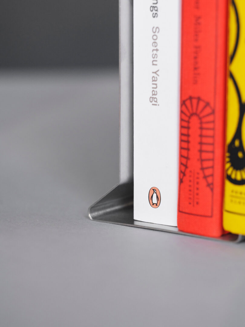 Primer plano de un sujetalibros de metal que sostiene tres libros en posición vertical, incluido un libro blanco con el logotipo de Penguin Books en el lomo.