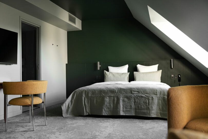 Moderna habitación de hotel con cama doble, paredes verdes, ropa de cama gris, techo inclinado, TV de pantalla plana y sillas de color amarillo mostaza. La luz natural entra a través de lucernarios.