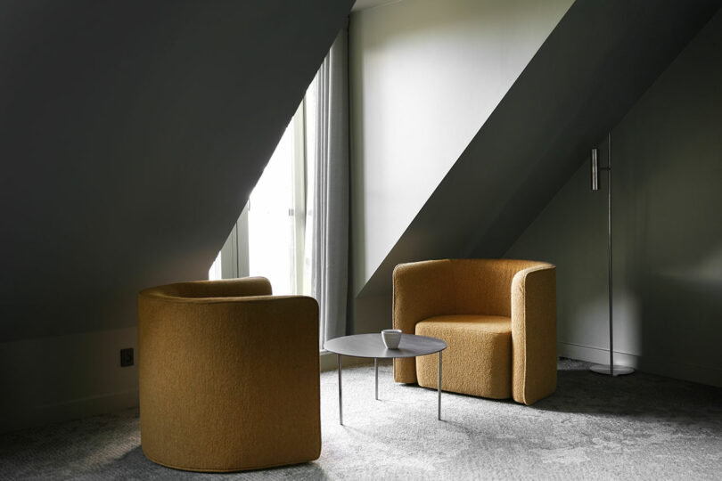 Dos sillones de color amarillo mostaza están frente a una mesa de café redonda negra con una taza encima, y la habitación tiene una alfombra gris, un techo inclinado, ventanas altas y una lámpara de pie.