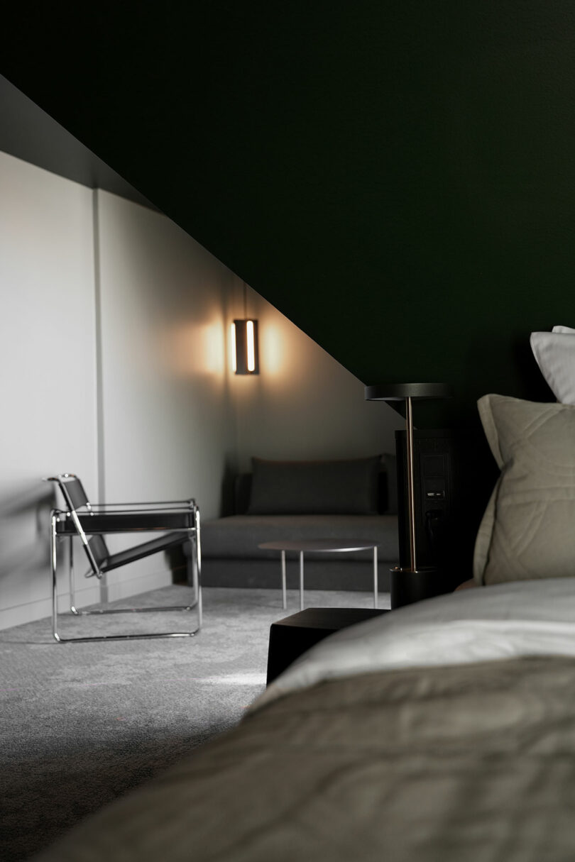 Dormitorio minimalista con una cama en primer plano y un sillón gris, un sofá, una mesa redonda y una lámpara de pared bajo un techo inclinado al fondo.