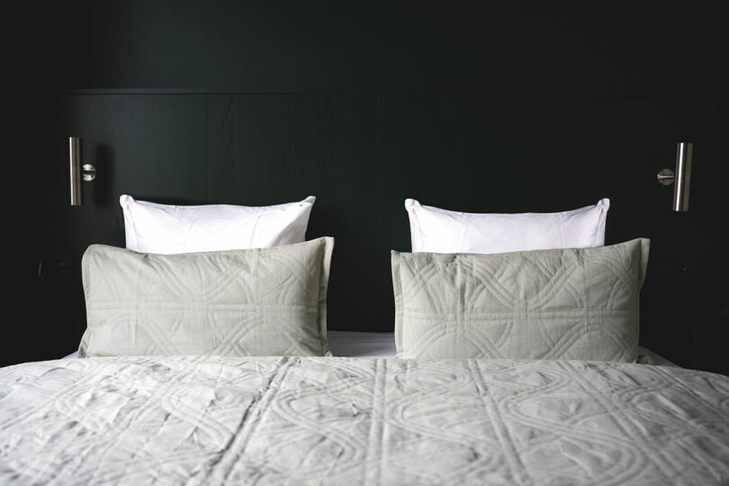 Una cama cuidadosamente hecha con dos almohadas blancas y dos almohadas acolchadas de color gris claro contra una pared oscura con dos luces de lectura montadas en la pared.