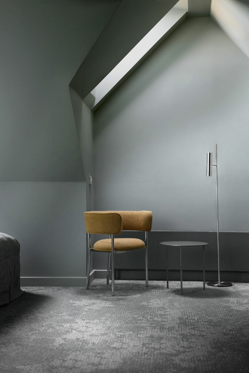 Es una habitación moderna y minimalista con techo inclinado y claraboya, dos sillas tapizadas en color canela, una pequeña mesa redonda, una lámpara de pie alta y una alfombra gris en el suelo.