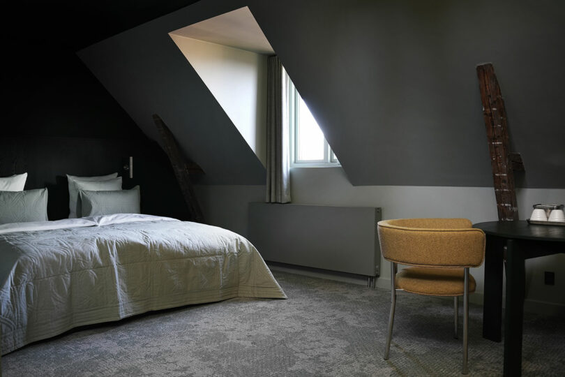 Moderna habitación de hotel con una cama grande, techo inclinado con claraboyas, vigas de madera vistas, una mesa redonda y una silla amarilla.