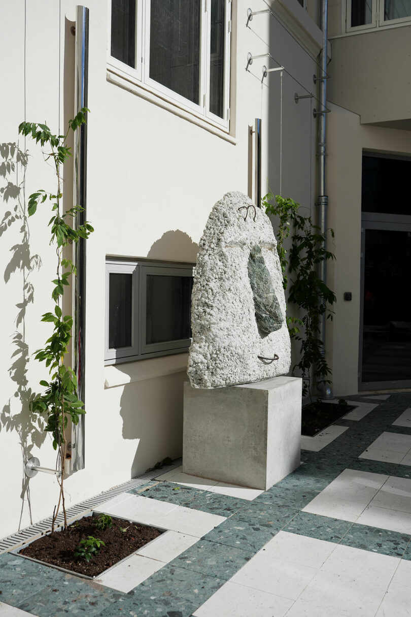 Una gran escultura de piedra irregular con un centro verde se alza sobre un pedestal de hormigón junto a un edificio de color claro, cerca de ventanas y plantas trepadoras jóvenes.