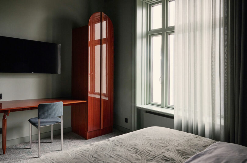 Un dormitorio sencillo con cama, escritorio y silla, televisor colgado en la pared, armario de madera y grandes ventanales con cortinas transparentes que dejan pasar la luz natural.