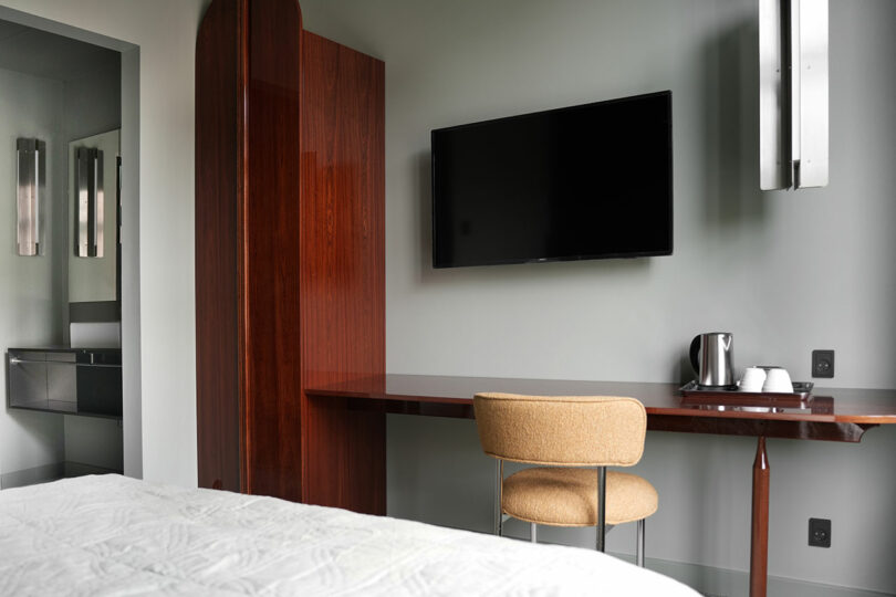 Una habitación de hotel con una cama, un televisor colgado en la pared, una mesa de madera con sillas y una bandeja con tetera y tazas; un baño visible a través de la puerta abierta.