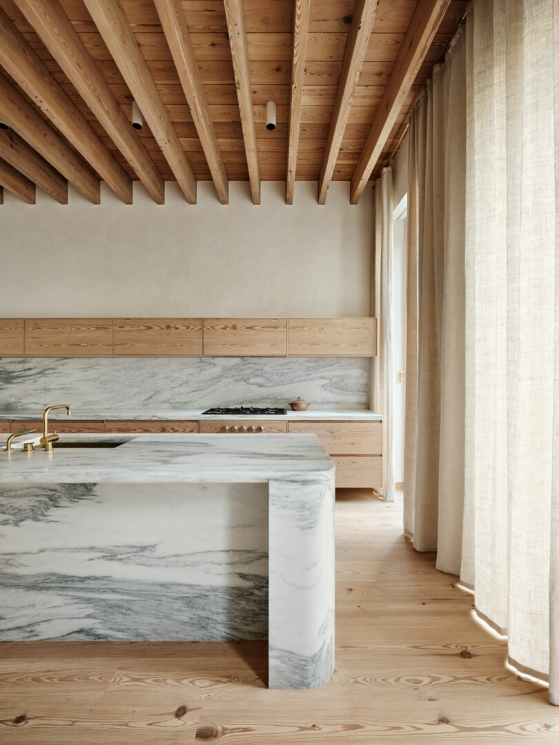 La moderna cocina de Brent Barker cuenta con vigas de madera a la vista en el techo, gabinetes de madera clara, una isla de mármol con protector contra salpicaduras a juego, grifos dorados y cortinas beige del piso al techo.