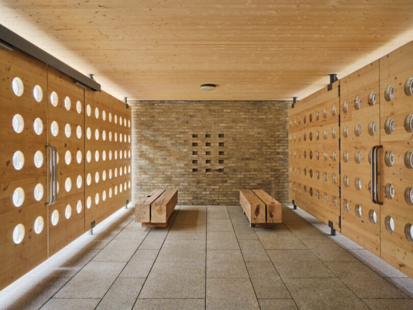 Habitación diseñada por Brent Buck con paredes y bancos de madera, cortes circulares en los extremos y paredes de ladrillo, suelo de baldosas y luces de techo.