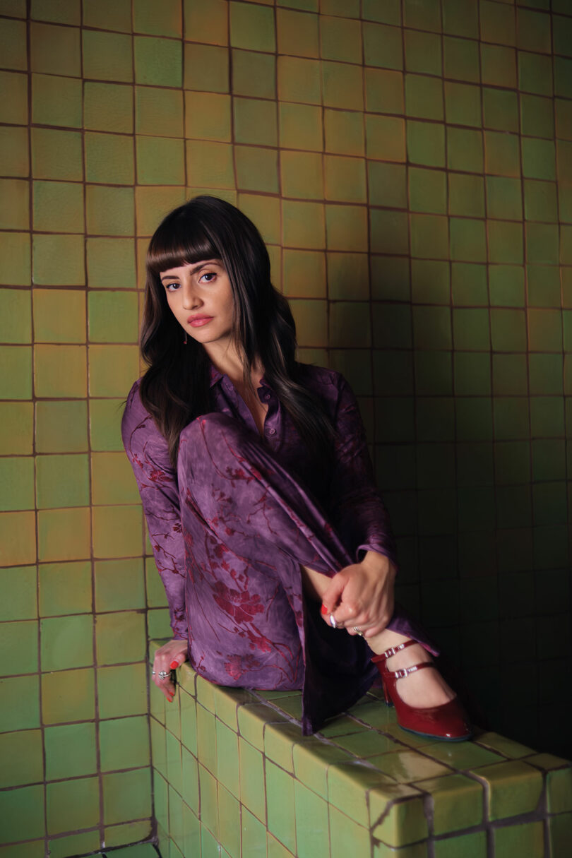 Maye Ruiz, una mujer con cabello largo y negro y flequillo, que lleva un vestido estampado de color púrpura y zapatos rojos, está sentada en una repisa de azulejos verdes y mira a la cámara.