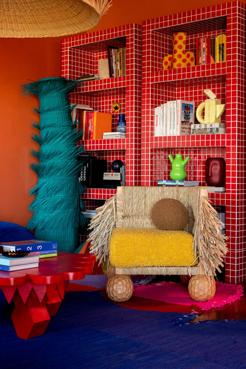 La sala ecléctica cuenta con estanterías de azulejos rojos, libros y decoración coloridos, sillas de jardín texturizadas, mesas angulares rojas y una escultura alta tejida en verde azulado en la pared naranja: un espacio de arte inspirado en Meyer Ruiz.