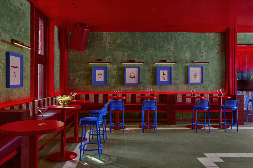 Bar moderno de Maye Ruiz con paredes y muebles de color rojo llamativo, paredes con textura verde, sillas azules, arte enmarcado y mesas con vasos.