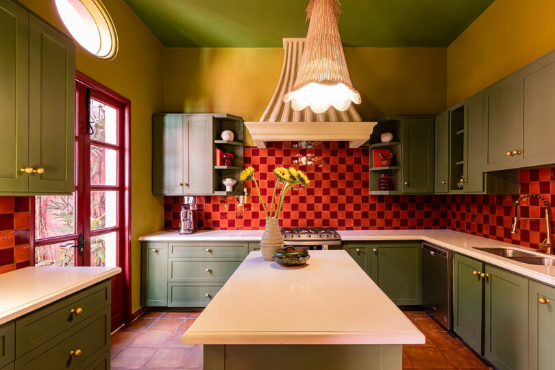 La cocina diseñada por Maye Ruiz cuenta con gabinetes verdes, un protector contra salpicaduras de cuadros rojos, encimeras blancas, una isla con un jarrón de girasol y una gran campana extractora blanca debajo de un techo verde.