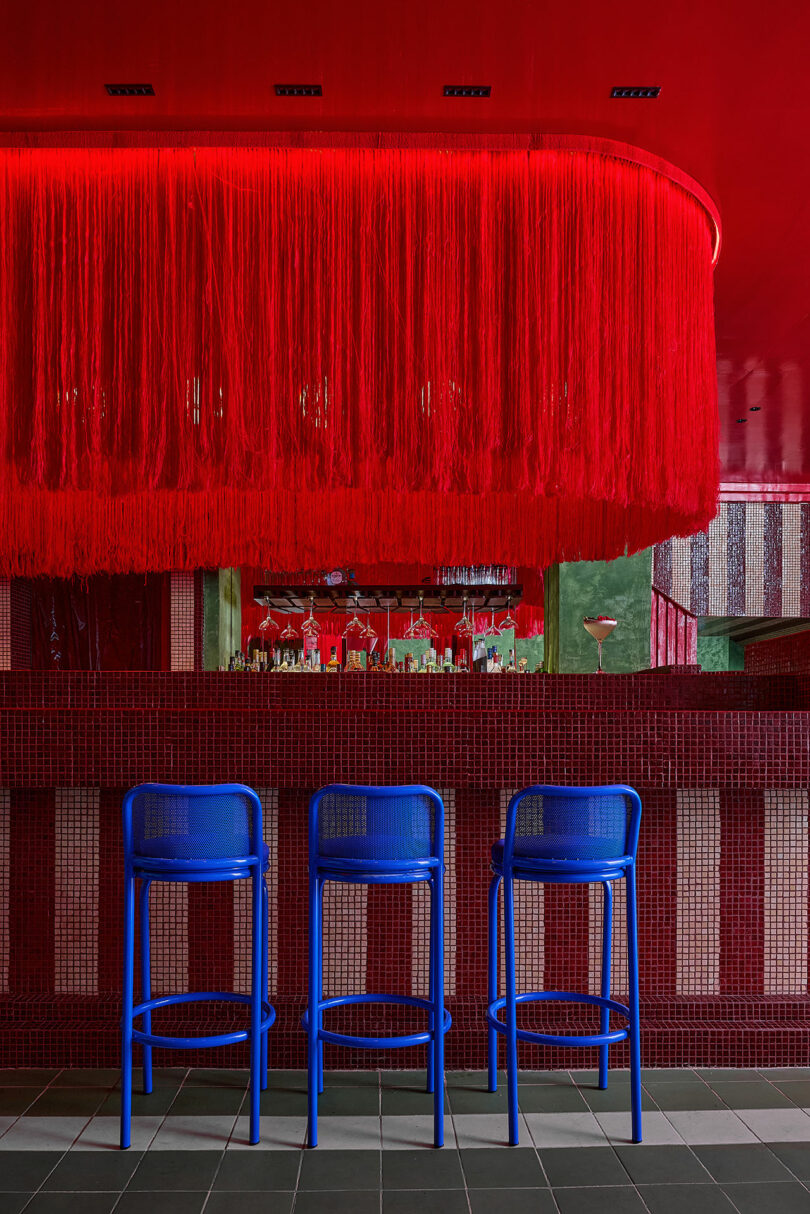El bar diseñado por Maye Ruiz cuenta con un mostrador de azulejos rojos, paredes con rayas verticales, tres taburetes azules y espectaculares detalles en el techo con flecos rojos.