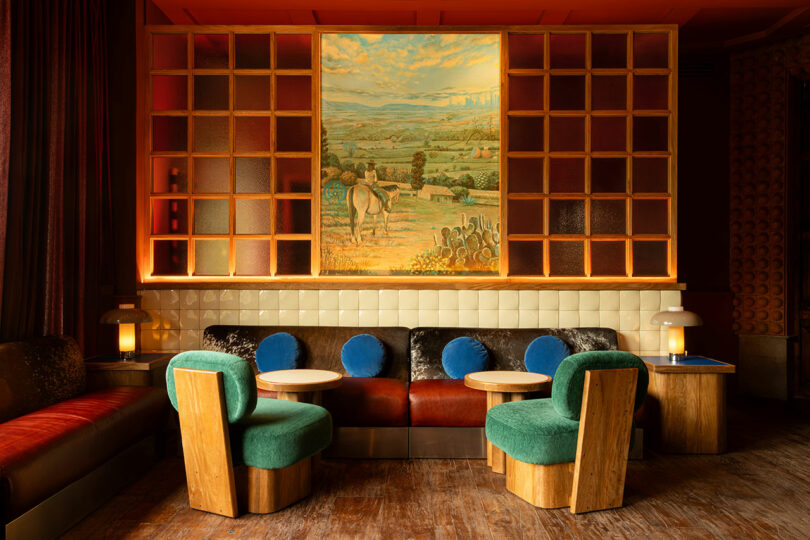 El acogedor salón cuenta con sillas verdes y azules, mesas redondas de madera, bancos tapizados y grandes pinturas de paisajes pastorales de Maye Ruiz en las paredes divisorias.