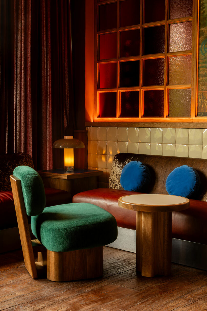 Una elegante zona de estar diseñada por Maye Ruiz cuenta con sillas tapizadas en verde, una mesa redonda de madera, un banco marrón con almohadas azules, lámparas de mesa y una cálida iluminación ambiental.