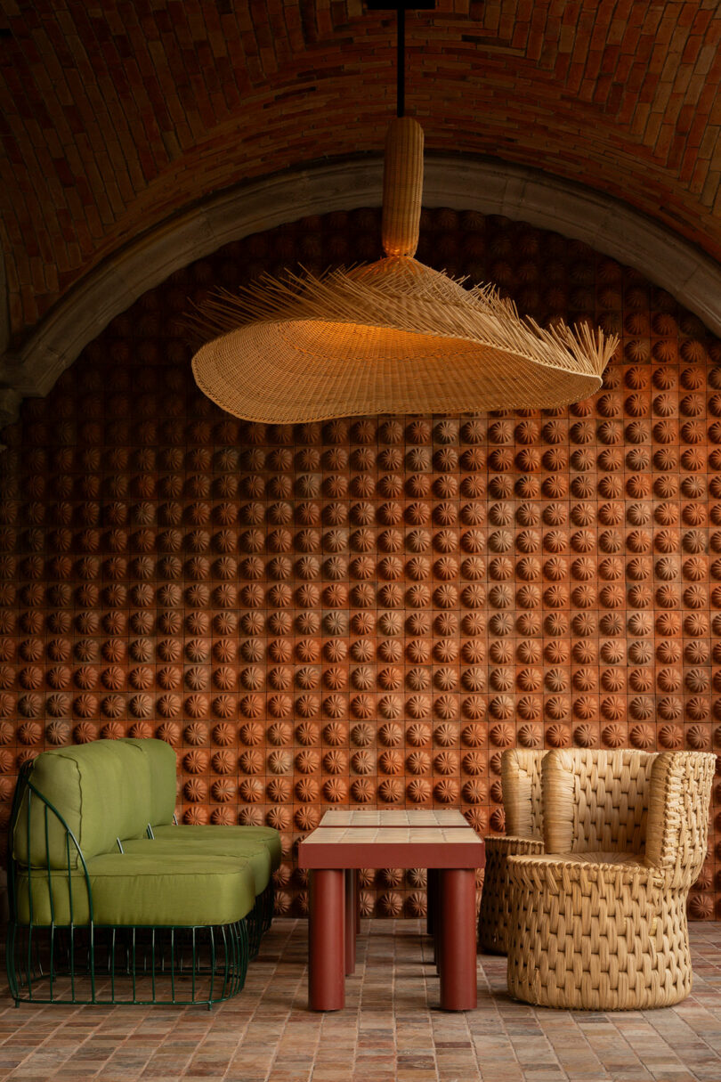El área del salón cuenta con un sofá verde con estructura de metal, sillas tejidas y una mesa rectangular roja debajo de una gran lámpara de araña tejida, contra una pared de ladrillos texturizados y un techo abovedado, diseñado por Maye Ruiz.