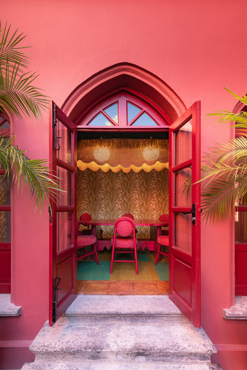 Puertas dobles rojas se abren a una habitación interior diseñada por Maye Ruiz, donde sillas rosas, una alfombra verde, una lámpara de araña, papel pintado estampado y dos palmeras enmarcan la entrada.