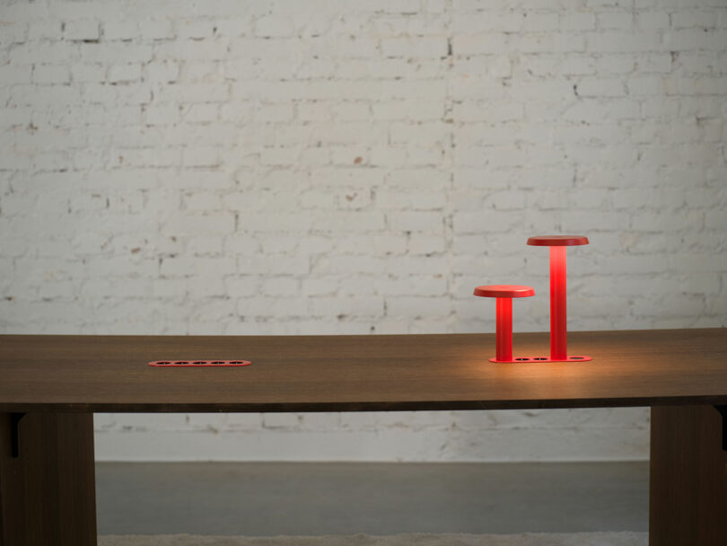 Una mesa de madera con dos modernas lámparas rojas en forma de seta y un juego de enchufes rojos descansa contra una pared de ladrillo blanco.