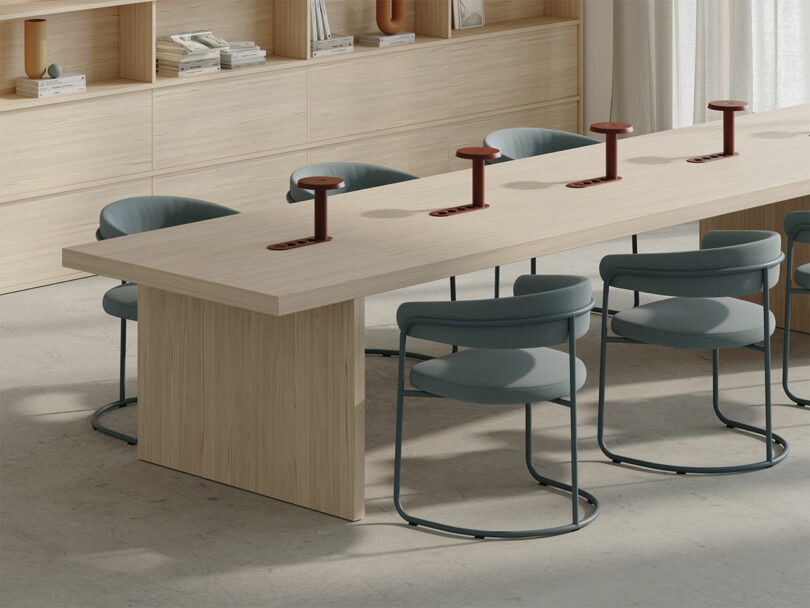 Moderna sala de conferencias con mesa de madera clara, sillas azules, estantes integrados y cuatro tomas de corriente rojas.