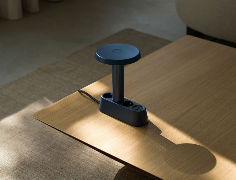 Hay una regleta de escritorio azul con dos tomas de corriente y dos puertos USB en la mesa de madera cerca del borde, con cables de alimentación a los lados.