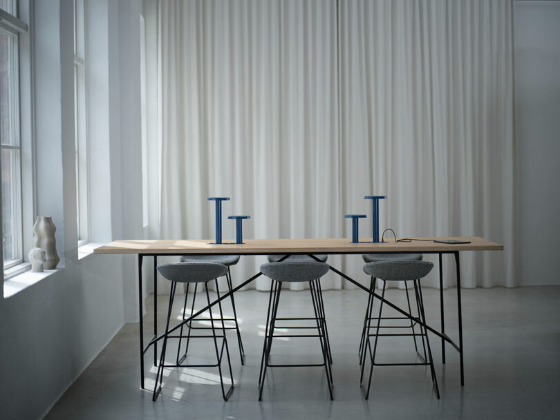 Sala de conferencias sencilla con una larga mesa de madera, cuatro taburetes grises, candelabros azules, un cuaderno y cortinas blancas que van desde el suelo hasta el techo.