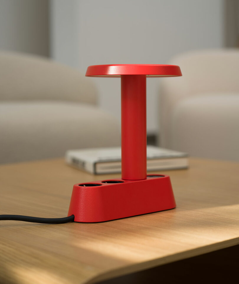 Una moderna lámpara de mesa roja con base rectangular se asienta sobre una mesa de madera con un libro borroso y una silla blanca al fondo.