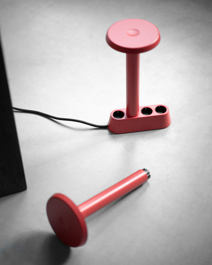 Dos objetos cilíndricos rojos, uno en posición vertical con tres enchufes y un cable de alimentación conectado, el otro en posición horizontal sobre una superficie gris claro.