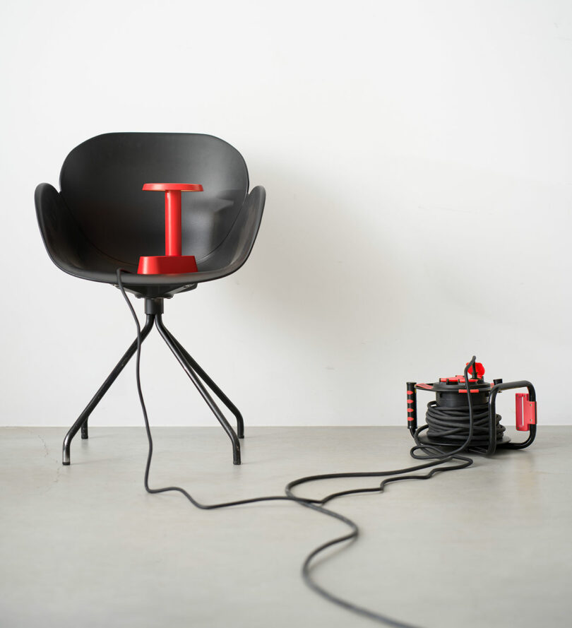 Una silla negra con una lámpara roja en el asiento junto a un carrete de cable de extensión negro y rojo sobre un piso claro con cables que se extienden hacia el primer plano.